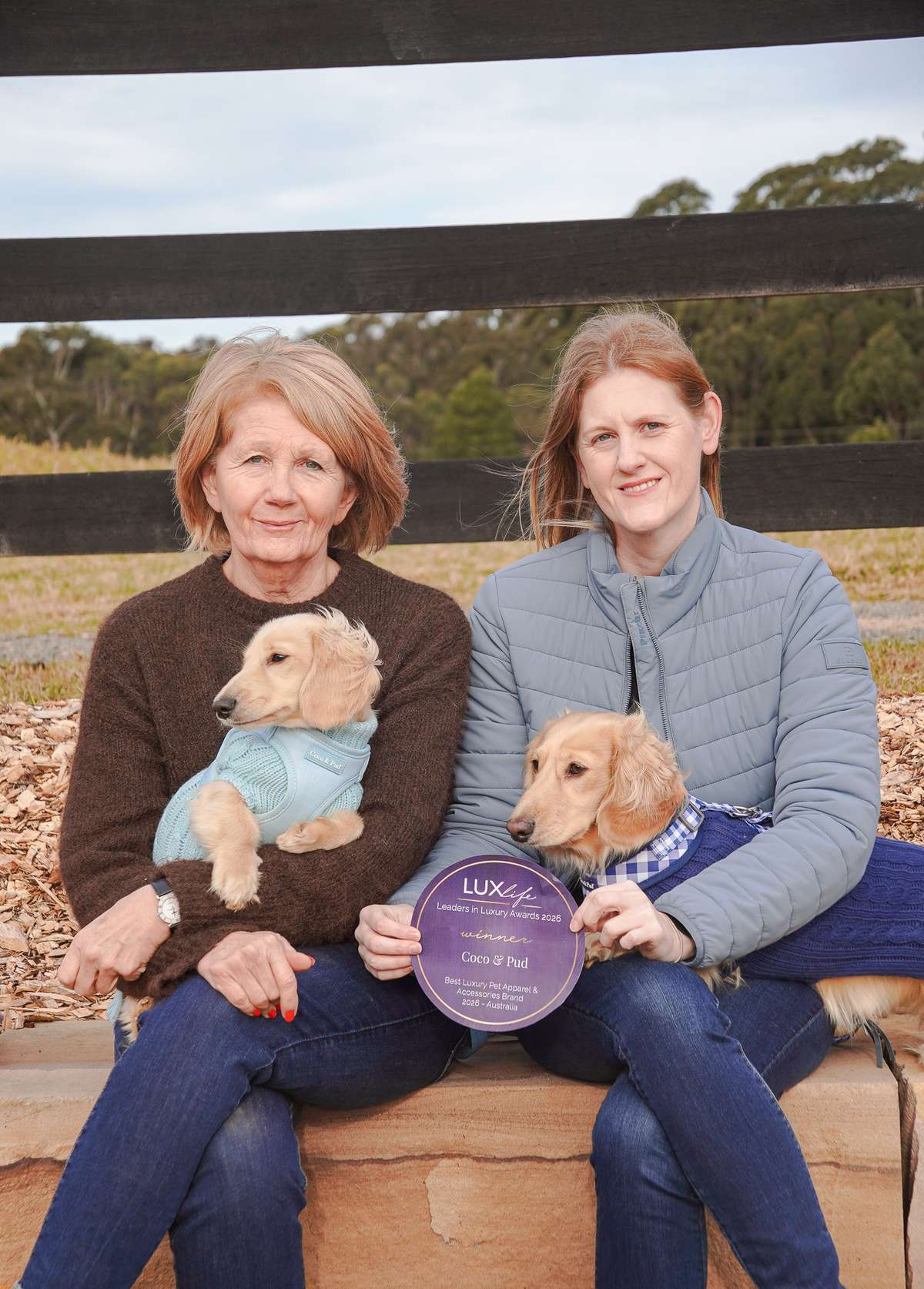 sarah and jane sitting outdoors holding small dachshunds in sweaters and a round purple award plaque.