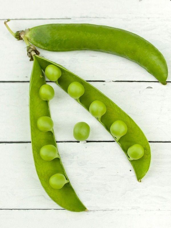 Two green pea pods, one open to reveal peas, on a white wooden surface.