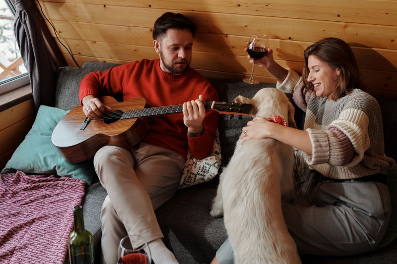 A couple relaxes with their dog as one person plays guitar and the other holds a glass of wine.