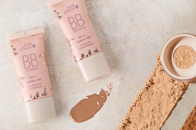 Two pink tubes of 100% Pure BB cream with a liquid swatch and powder on a light surface.