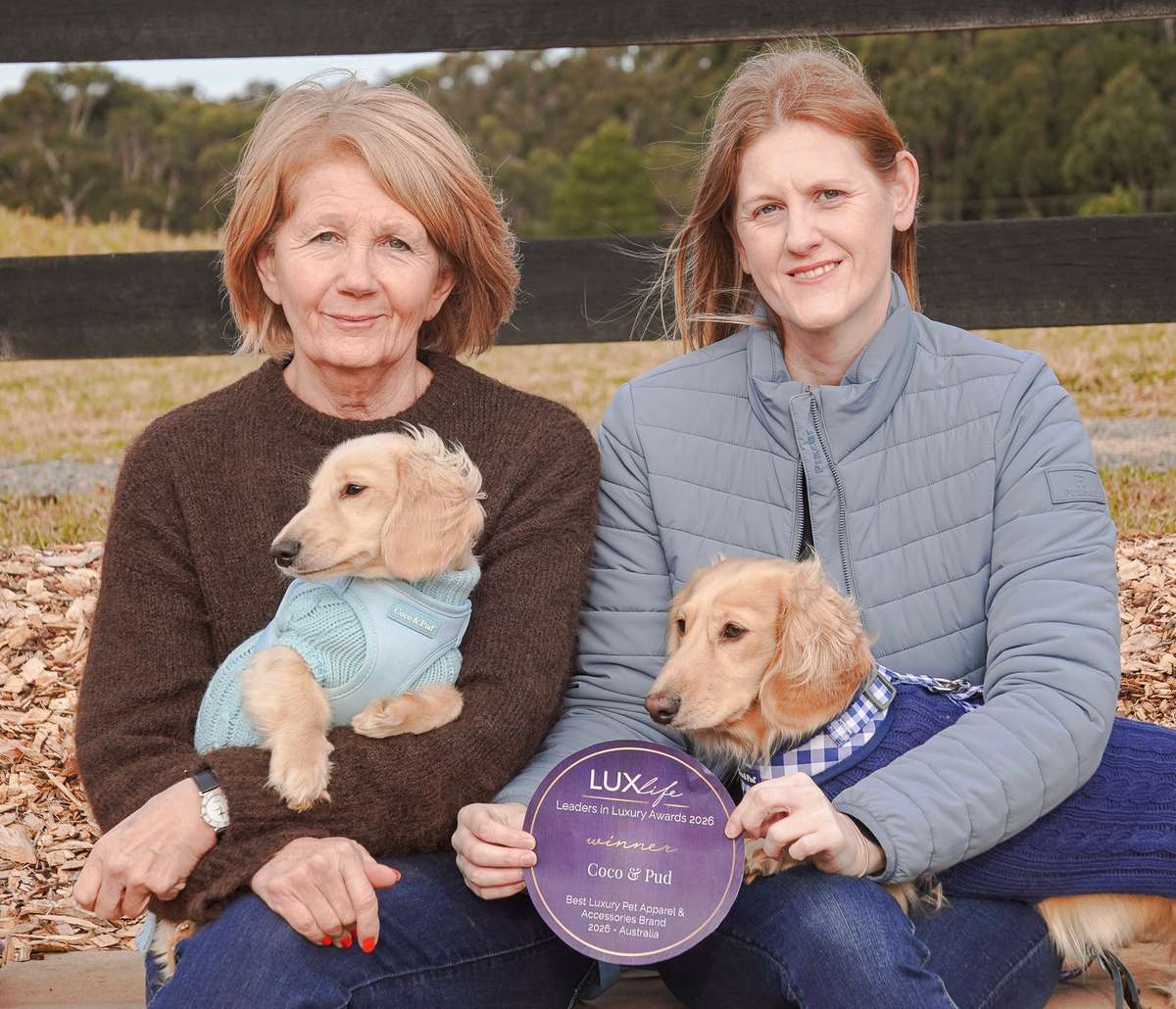 sarah and jane holding small dachshunds in sweaters display a round purple award plaque outdoors.