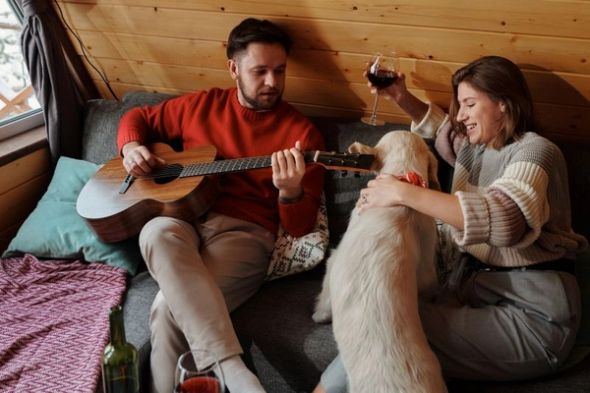 A couple relaxes with their dog as one person plays guitar and the other holds a glass of wine.