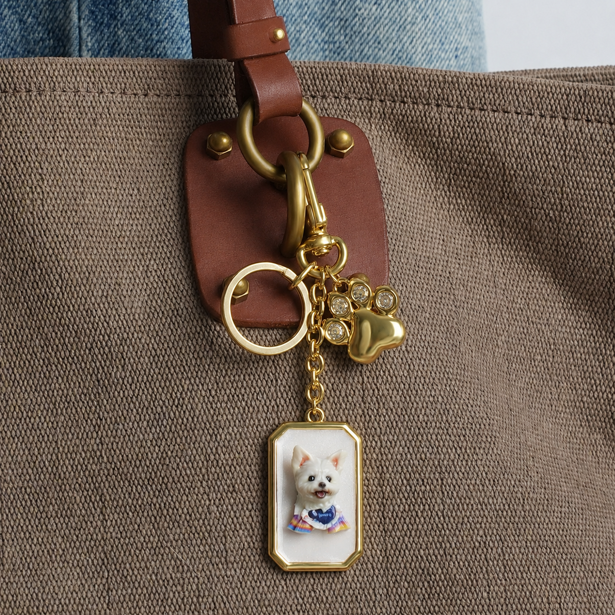Gold keychain with a 3D white dog portrait and paw charm clipped to a brown canvas tote bag.