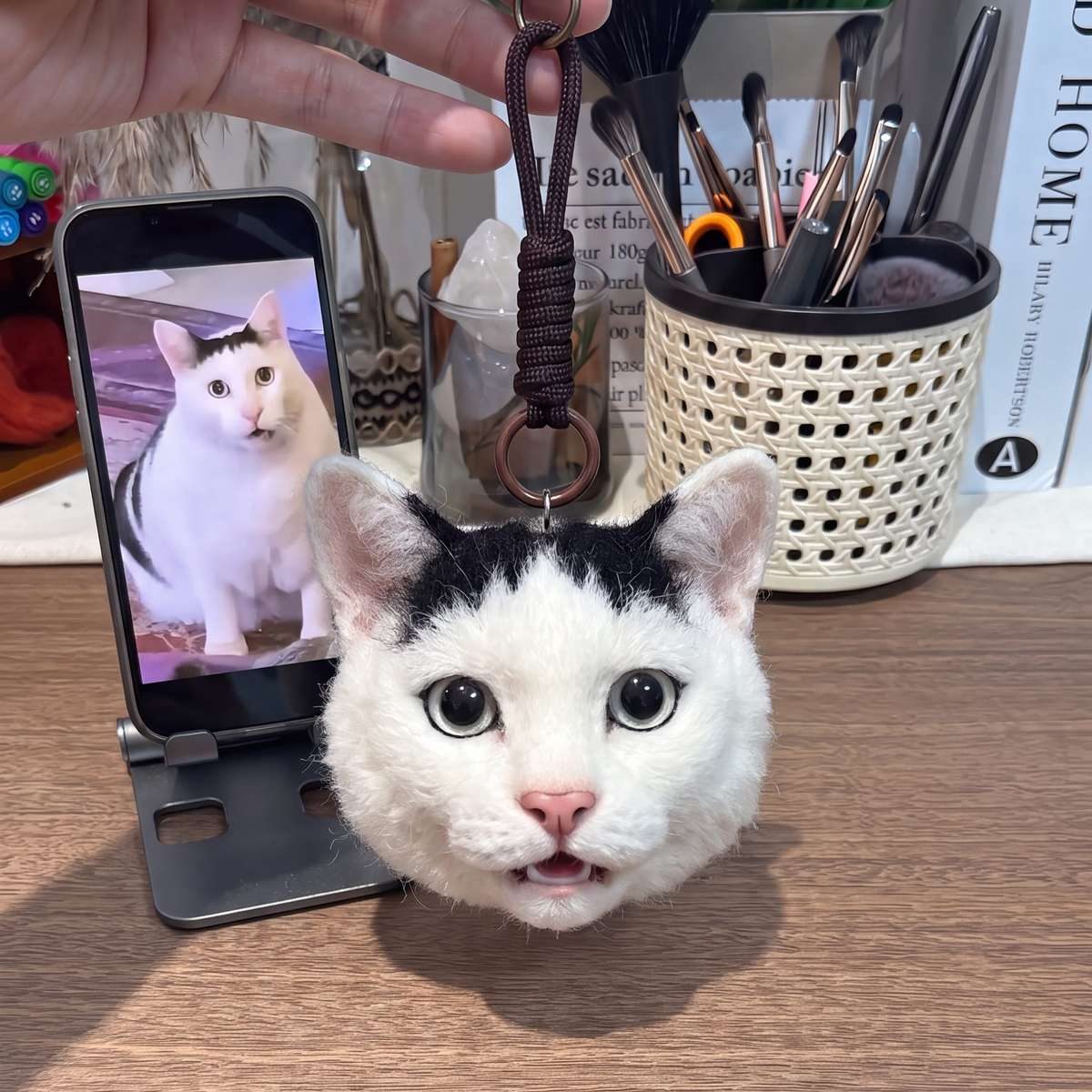 A needle-felted cat head keychain held beside a phone displaying a photo of the real cat.