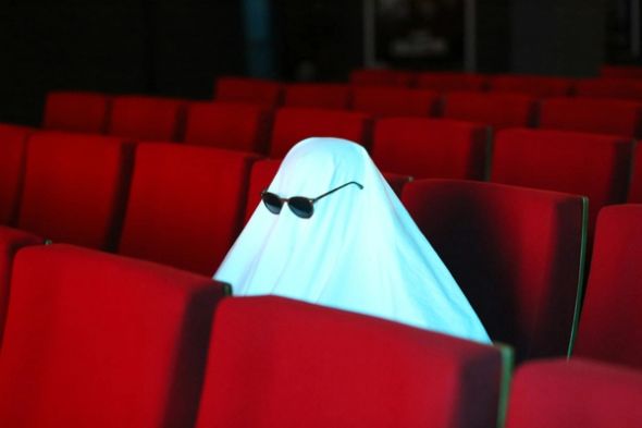 Ghost wearing sunglasses sits among red cinema seats.