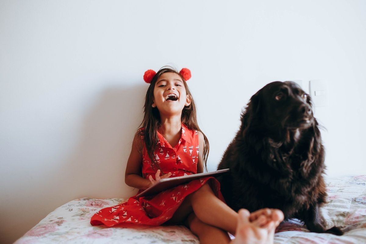 A young girl in a red dress laughs while holding a tablet beside a fluffy black dog on a bed.