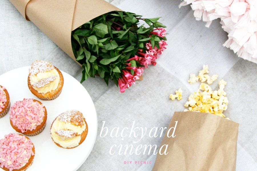 Mother's Day backyard cinema picnic setup with pink cupcakes, fresh popcorn, and a flower bouquet on a blanket.
