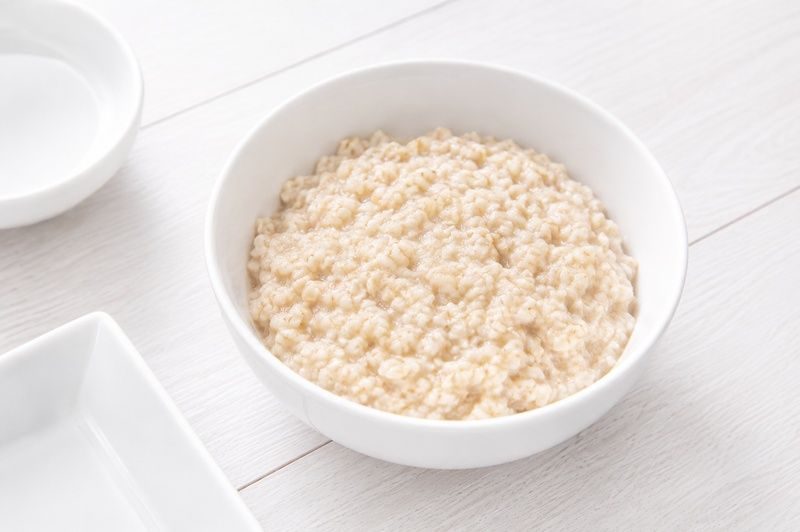 A white bowl filled with plain cooked oatmeal on a light wooden surface.
