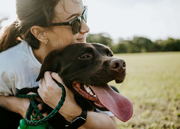 A woman wearing sunglasses smiles and hugs a panting brown dog in a grassy field.