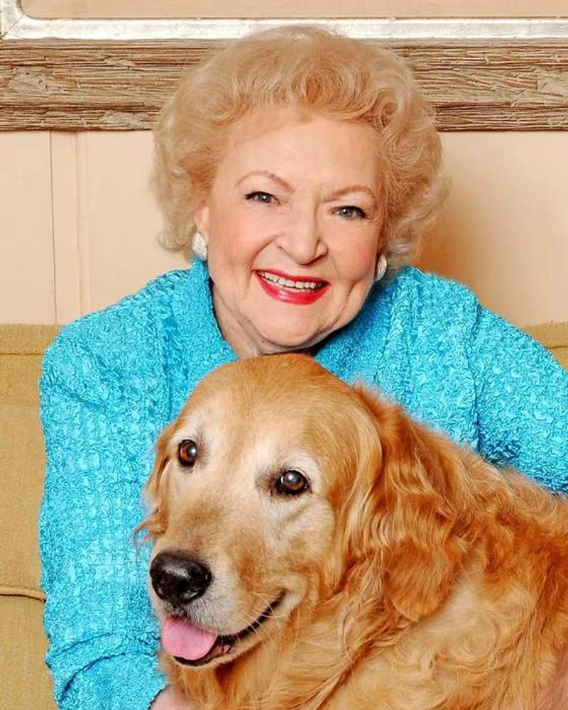 Betty White wearing a bright blue jacket smiles while posing with a Golden Retriever.