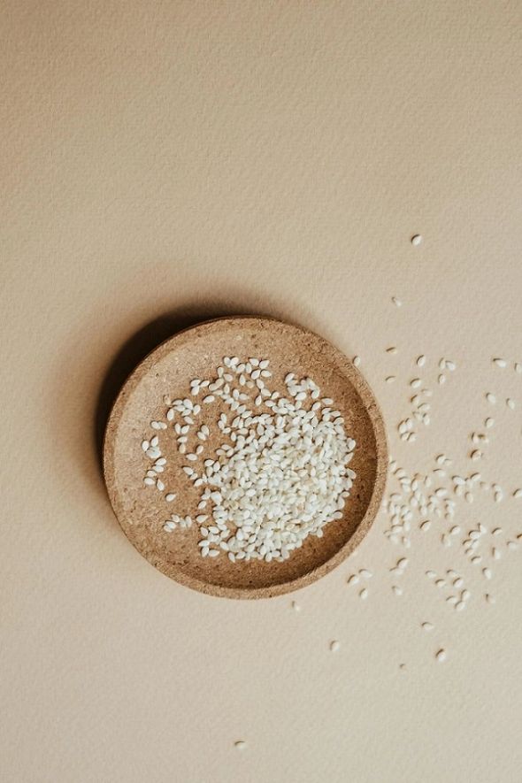 A wooden bowl filled with white sesame seeds, with more seeds scattered across a beige surface.