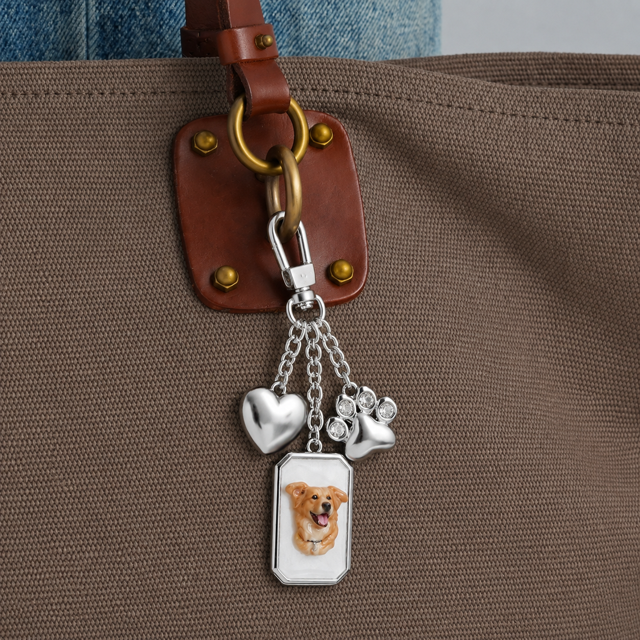 A silver bag charm with a heart, jeweled paw print, and 3D dog portrait attached to a brown tote bag.