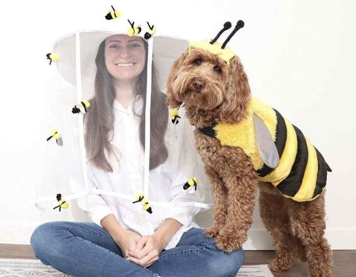 Woman in a beekeeper costume sits beside a dog dressed as a bee, with small bees around them.
