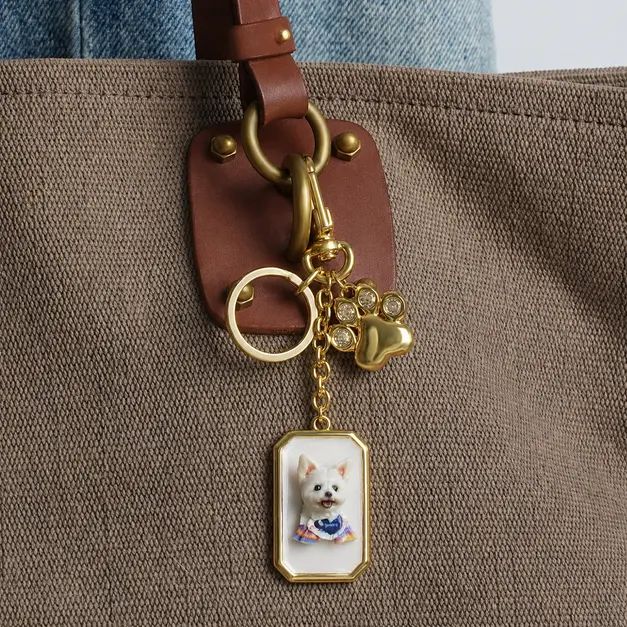 Gold keychain with a 3D white dog portrait and paw charm clipped to a brown canvas tote bag.