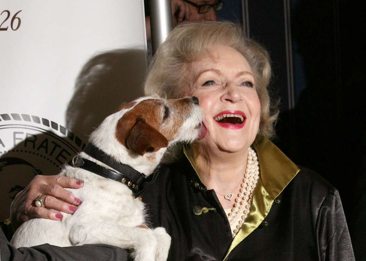 Betty White smiles happily as a small brown and white dog licks her cheek.