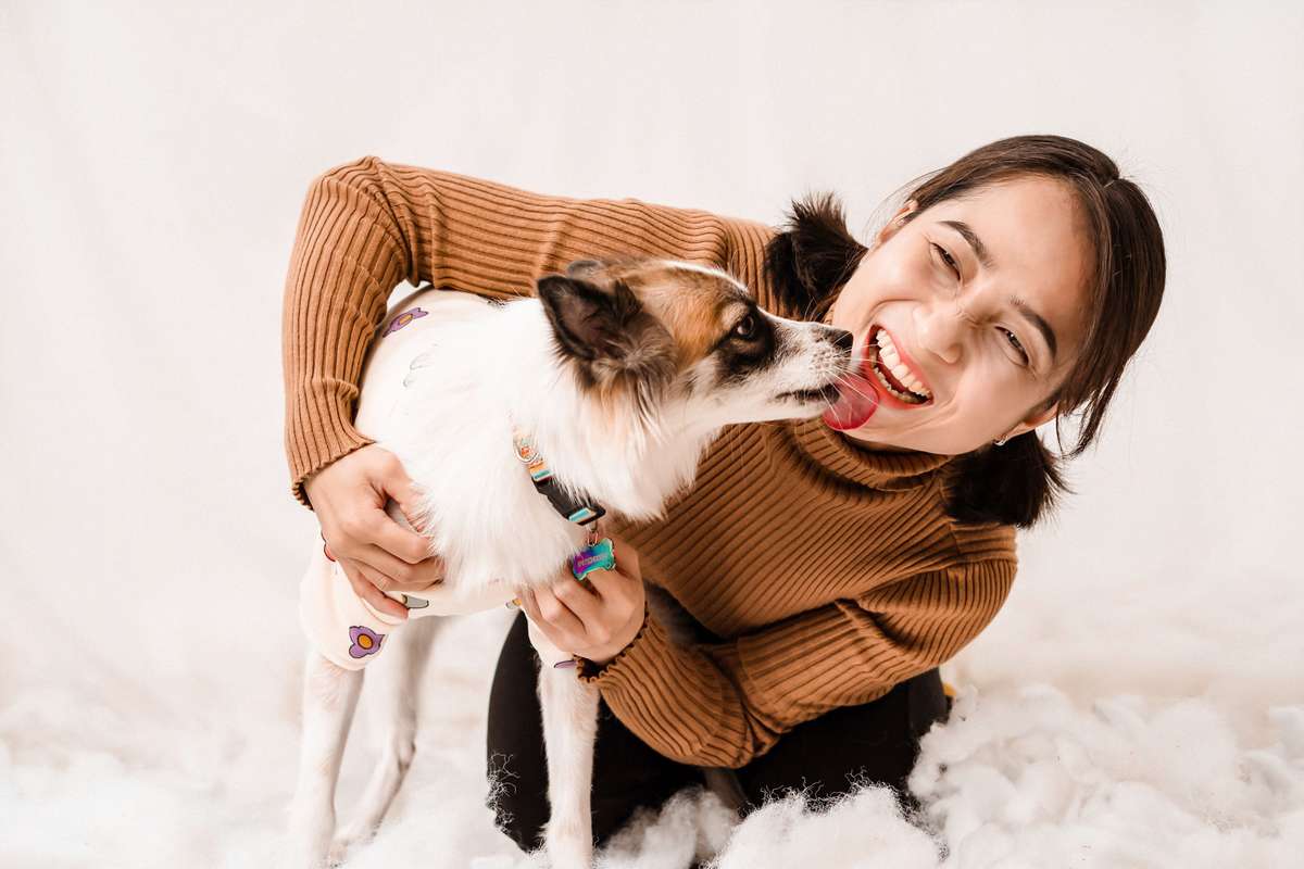 A smiling woman in a brown sweater holds a small dog licking her cheek.