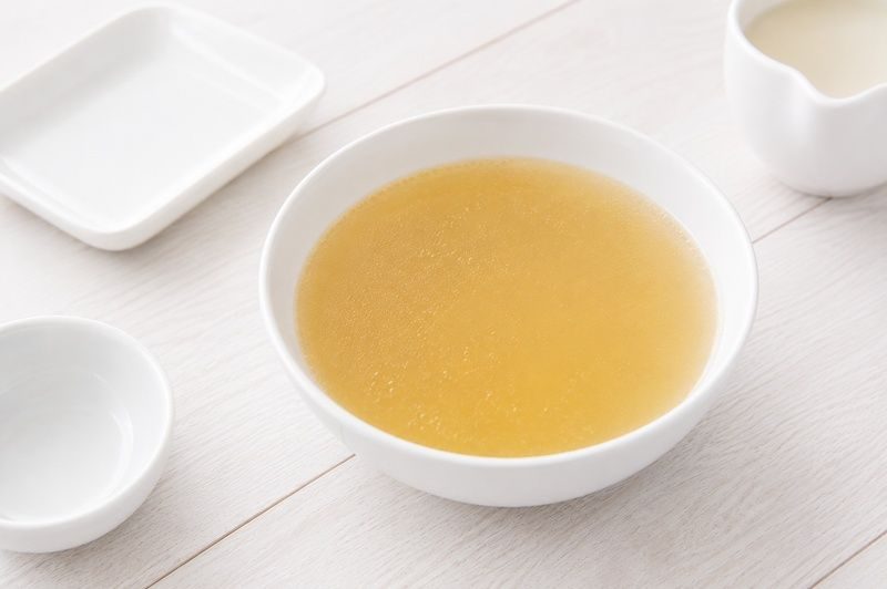 A white bowl of bone broth on a light wooden table surrounded by small white dishes.