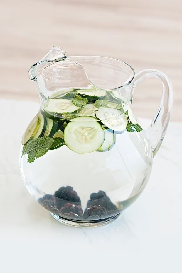 A glass pitcher filled with water, sliced cucumbers, mint leaves, and blackberries.