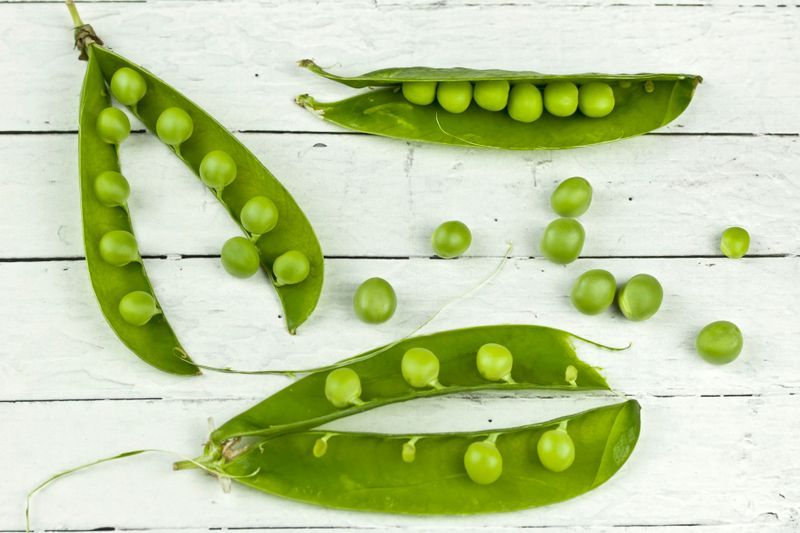 Open pea pods and loose green peas arranged on a white wooden surface.