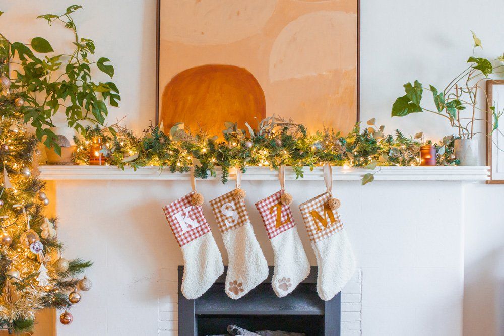 Personalized no-sew DIY dog stockings with paw prints hanging on a fireplace mantel with Christmas greenery and lights.