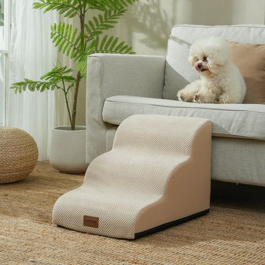 A fluffy white dog sits on a grey sofa beside beige foam pet stairs.