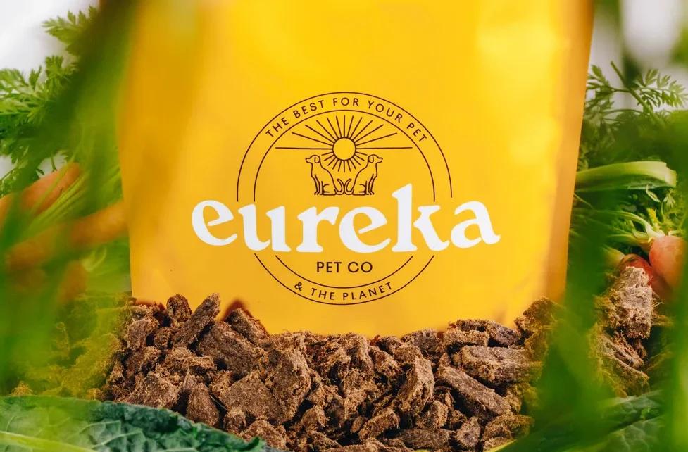 Eureka Pet Co yellow bag of Australian air dried dog food surrounded by fresh vegetables like carrots and kale.